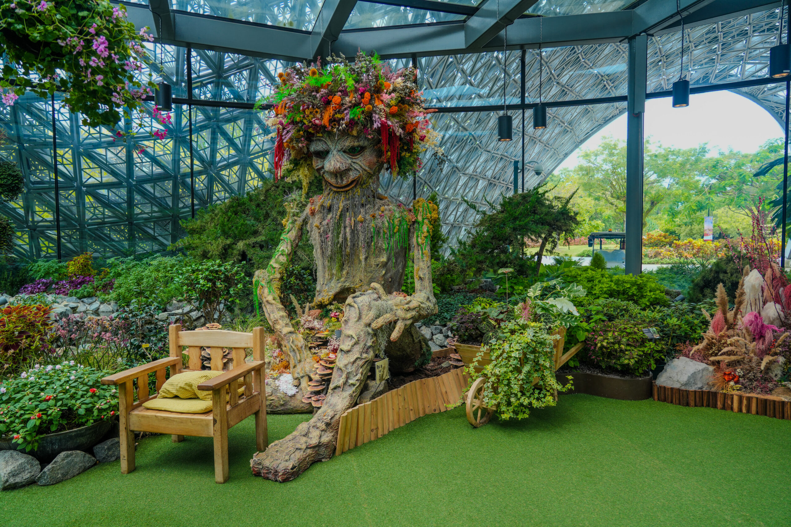 A whimsical indoor garden scene featuring a large tree-like sculpture with arms and a floral crown, surrounded by lush greenery, flowers, and a wooden bench in a glass atrium.