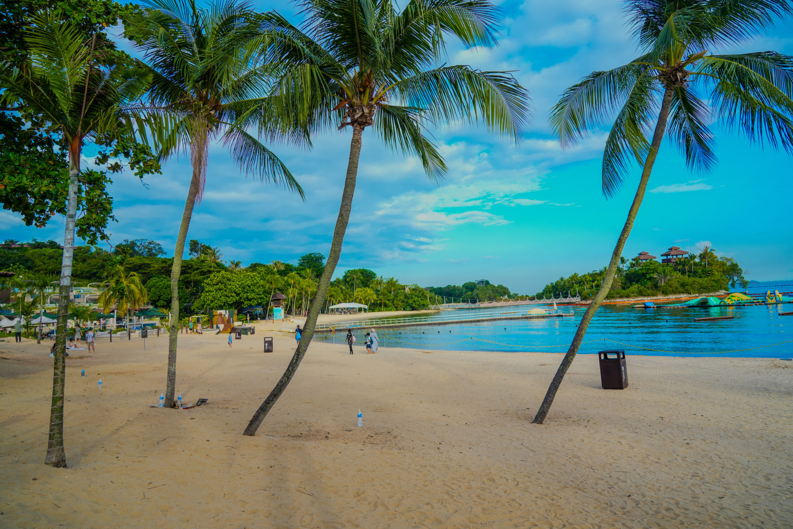 A peaceful tropical beach scene with golden sand, gently swaying palm trees, calm blue water, and a few people walking along Siloso Beach in Singapore.