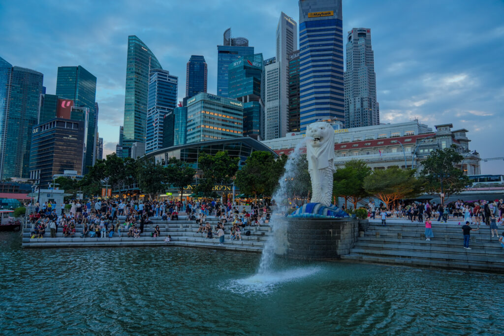 Singapore’s famous Merlion statue spouting water with a bustling crowd in front, and the modern skyline of downtown Singapore in the background during dusk.