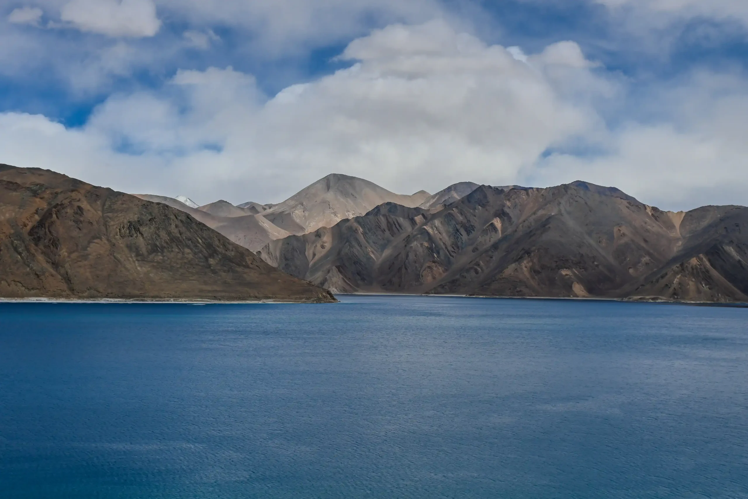 Bright blue Pangong Lake contrasting with barren brown mountains under a dramatic sky in Leh, Ladakh.