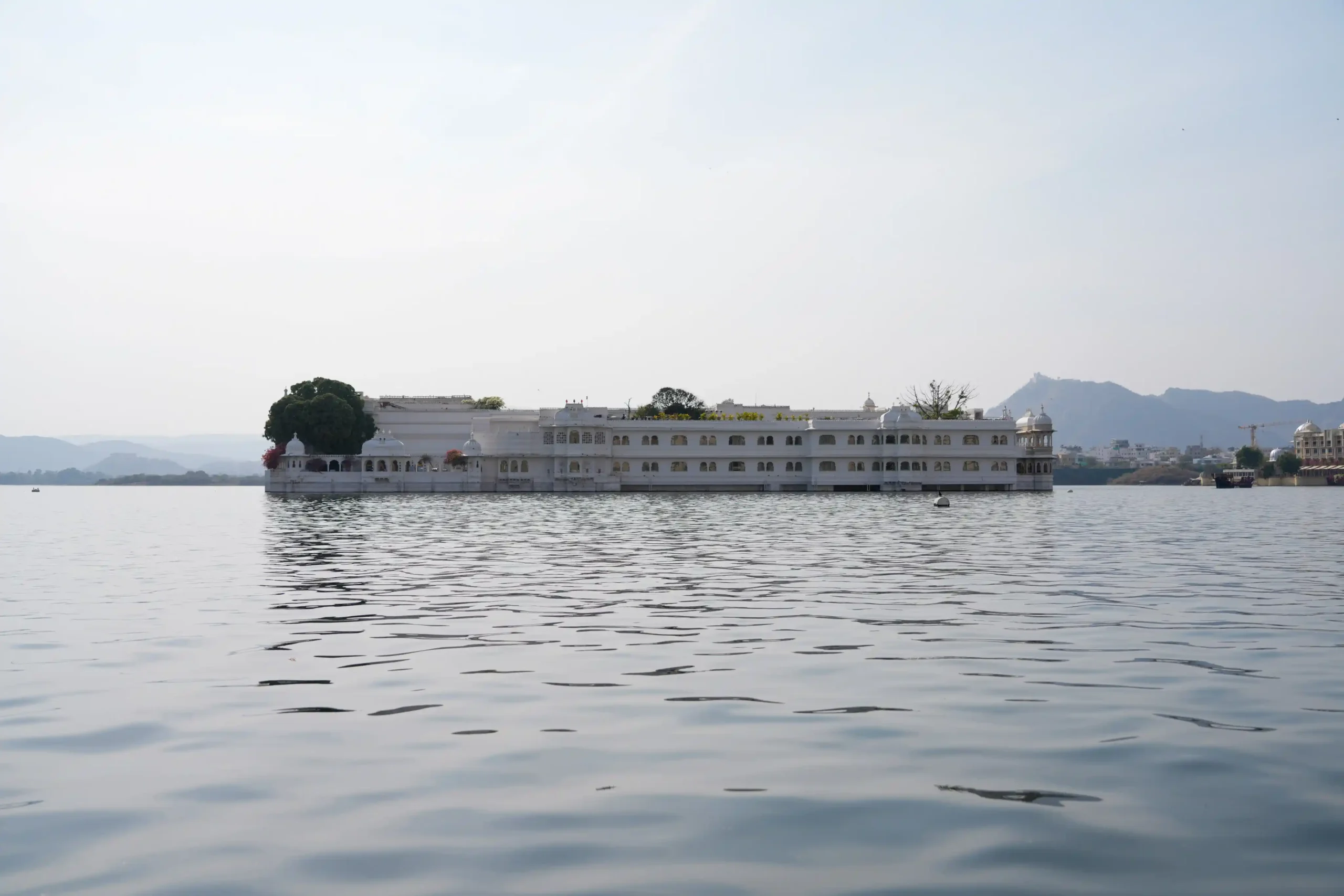 White marble Taj Lake Palace floating on Lake Pichola, Udaipur, with Aravalli hills in the background.