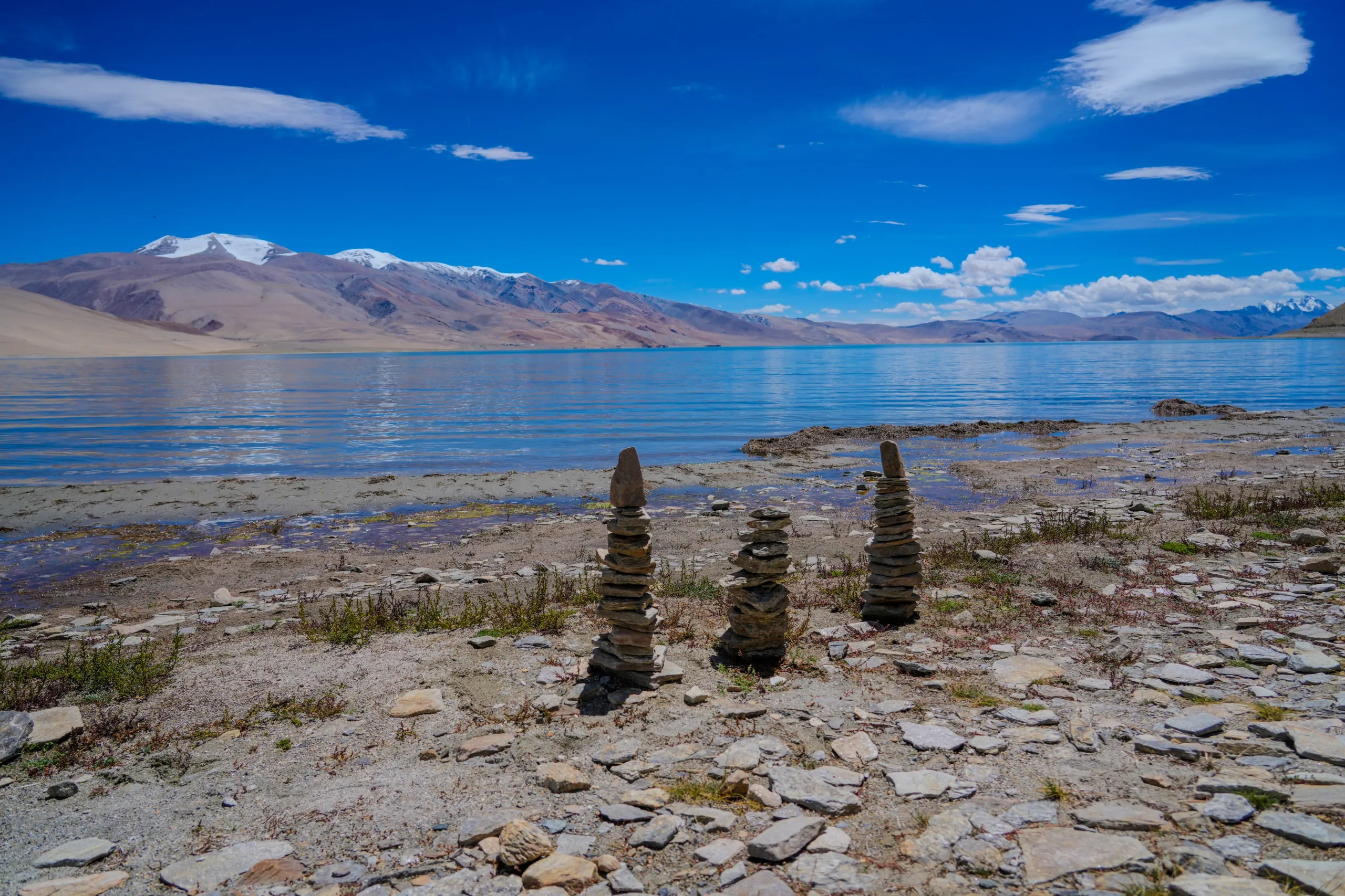 Stone cairns on the shore of Tso Moriri Lake with snowy peaks and blue sky in Ladakh